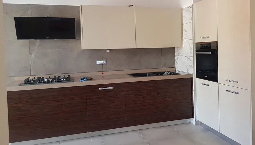 Cucina lineare con basi in legno scuro venato, pensili color crema e colonne con forno incassato